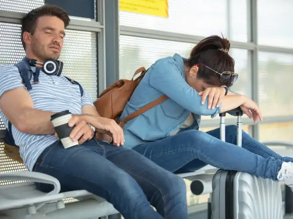 Erschöpftes Paar wartet auf den Flug. Der Mann schläft mit Kaffee, die Frau hat den Kopf auf den Koffer gelegt. Der Jetlag geht nicht weg.