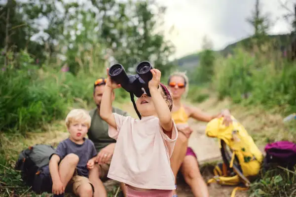 Familie im Urlaub mit Kleinkind: Ein Mädchen schaut durch ein Fernglas, während ihr Bruder und die Eltern im Hintergrund sitzen.
