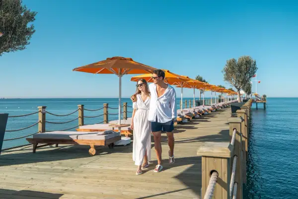 Ein Paar spaziert über einen Holzsteg am Meer, gesäumt von Liegen und Sonnenschirmen. Ein perfekter Urlaub im Arum Barut Collection.