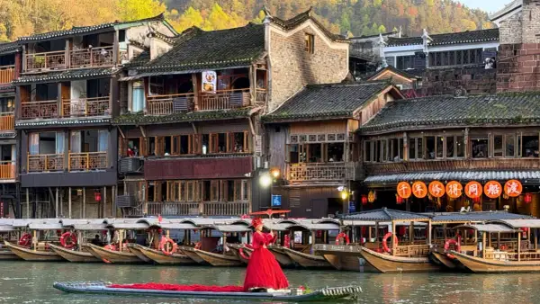 Ein Urlaub in China: Frau in rotem Kleid auf Boot vor traditionellen Häusern und Booten am Fluss.