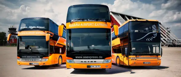 Drei orange-schwarze Doppeldeckerbusse von Allgäu Airport Express stehen bereit. Informieren Sie sich über die alex airport shuttle preise für Ihre Reise.
