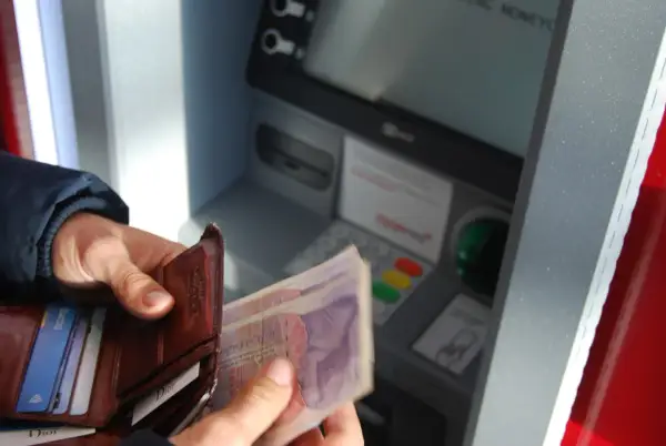 Person hält Geld und Portemonnaie vor Geldautomat. Hier kann man bei der Bank Geld wechseln.