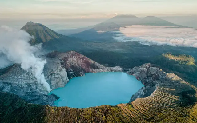 Der Ijen-Krater auf der **Java Insel** mit seinem türkisfarbenen Säuresee und rauchenden Vulkanflanken, umgeben von grünen Bergen und Wolken.