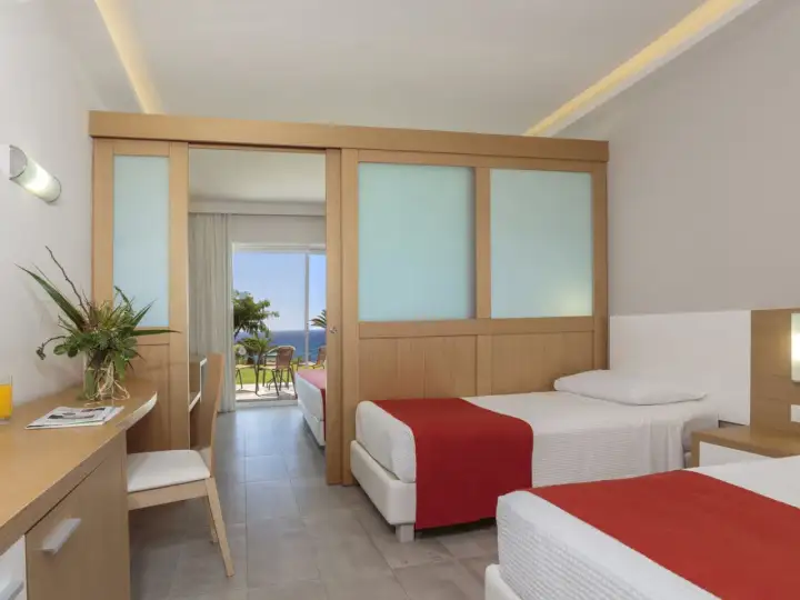 Zwei Betten mit roten Decken im Rodos Princess Beach Hotel. Ein Schreibtisch mit Blumen und Blick aufs Meer.